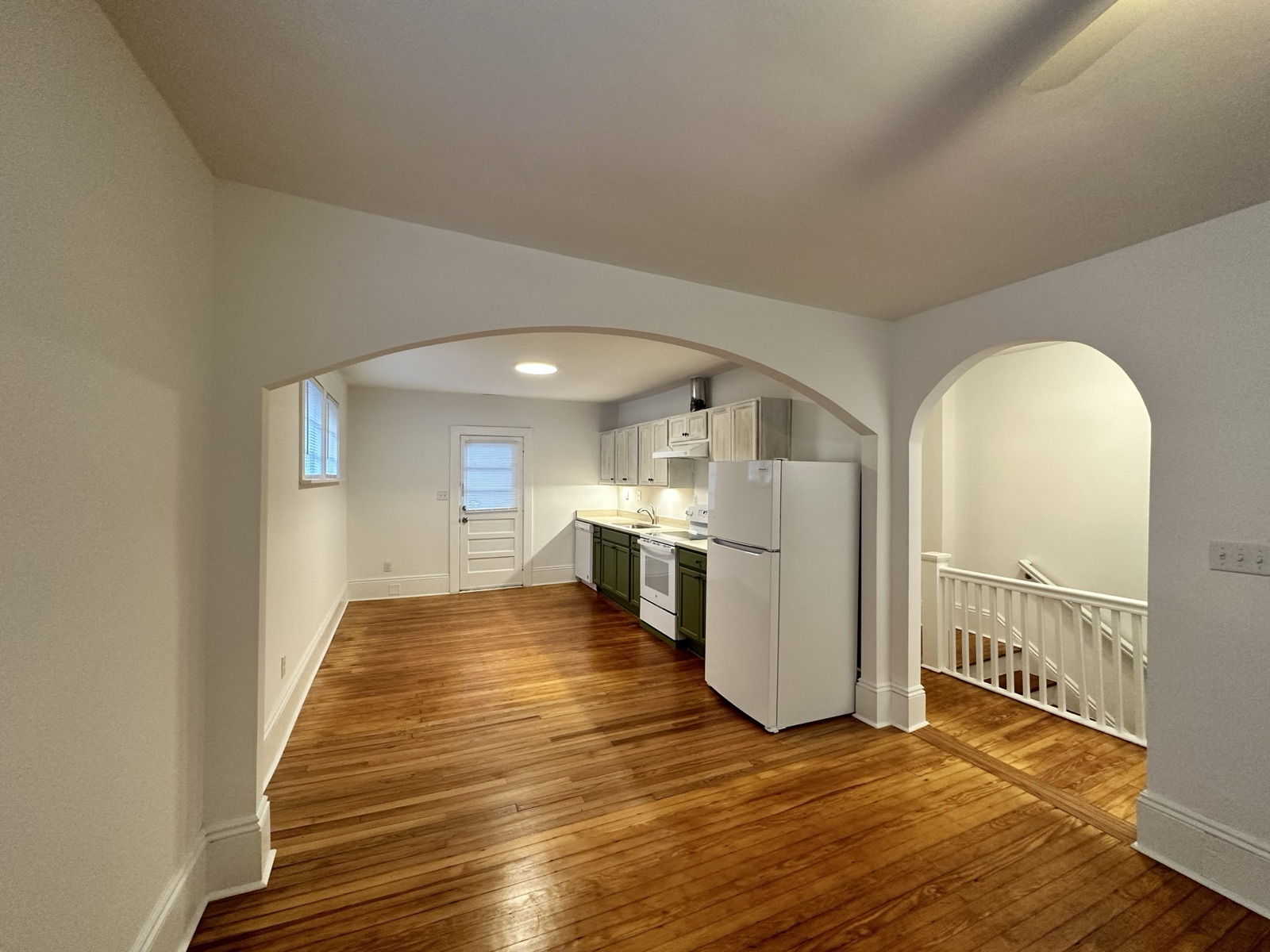 Kitchen with sage-green lower cabinets, whitewashed uppers, white appliances, and heart-pine floors flowing through an archway to the living area